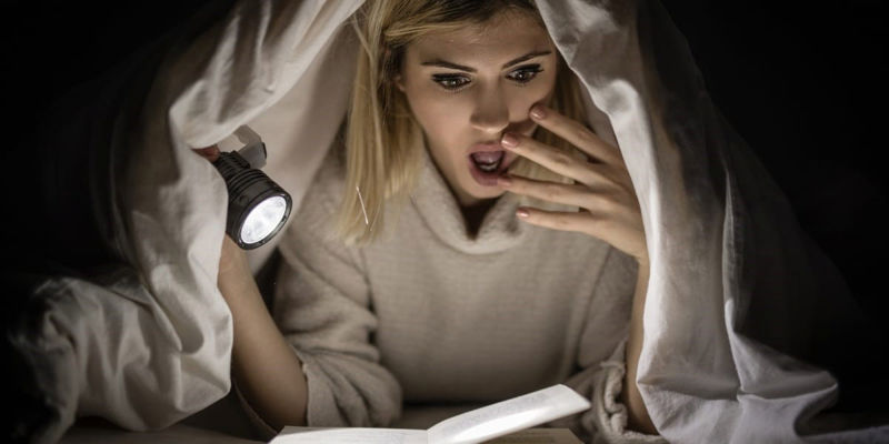 زنی با چراغ قوه در حال خواندن کتاب‌های ترسناک است