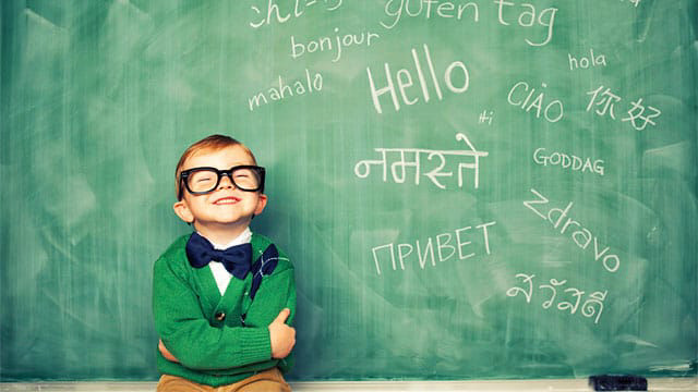 تصویر یک پسر بچه با عینکی بزرگ و خندان جلوی تخته سیاهی که به زبان‌های مختلف بر روی آن سلام نوشته شده است