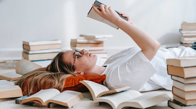 دختری عینکی که دراز کشیده و در میان انبوه کتاب‌ها کتاب می‌خواند