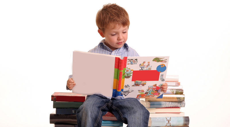 پسر بچه‌ای که بر روی کتاب‌ها نشسته و کتاب کودکان می‌خواند