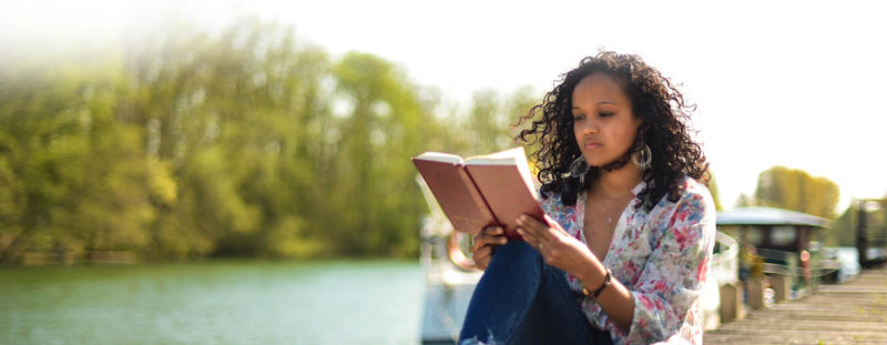 دختری که کنار دریاچه نشسته و کتاب می‌خواند