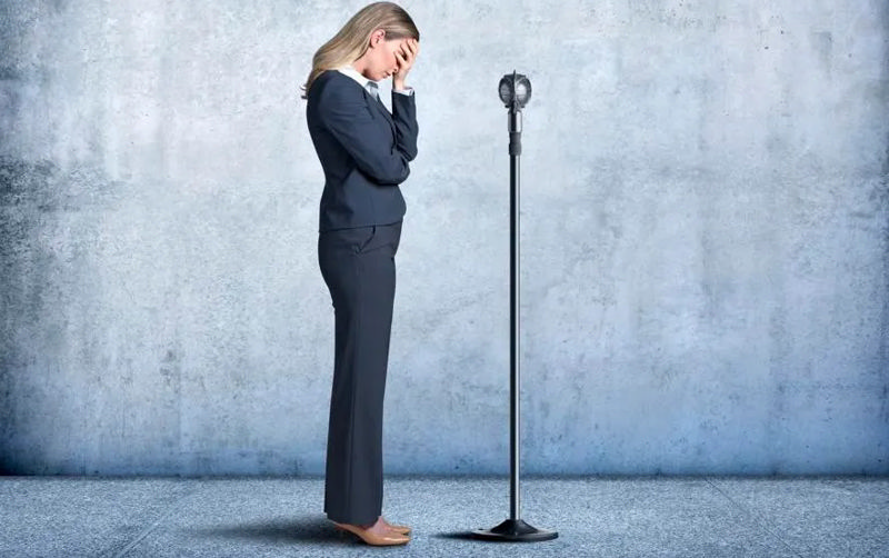 زنی که جلوی میکروفون ایستاده و به‌خاطر ترس از سخنرانی سرش را به دستش تکیه داده است