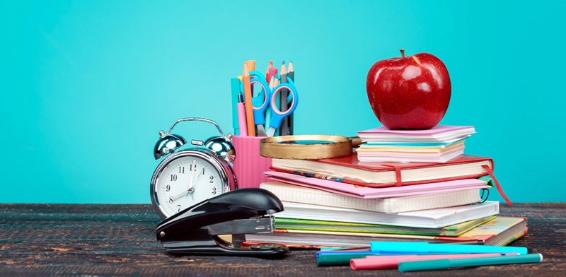 چند دفتر و کتاب بر روی هم و یک سیب روی آن‌ها قرار داده شده است. یک ساعت و منگنه و جامدادی در کنار کتاب‌ها قرار دارند.