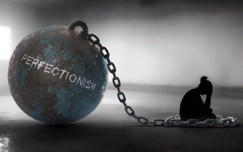 یک توپ فلزی سنگین با واژه کمالگرایی که انگار مردی را اسیر کرده است